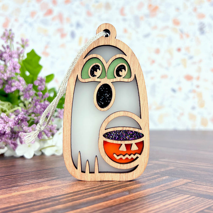 The Halloween Trick-or-Treating Ghost Ornament, 3.8" by Forged Flare® is a delightful wooden decoration featuring a ghost holding a pumpkin candy basket. Accented with glittery black and orange details, the ghost has green eyes and a surprised expression. The background showcases a wooden surface and blurred flowers, enhancing the charm of this Halloween piece.