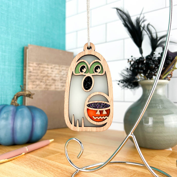 The Halloween Trick-or-Treating Ghost Ornament by Forged Flare® is an adorable wooden decoration measuring 3.8 inches and hangs from a string. The ghost has green eyes and a black mouth, holding a small orange pumpkin with a glittery purple interior. In the background, a blue pumpkin, an open book, a pink pen, and a ceramic vase with dark feathers add to the seasonal charm.