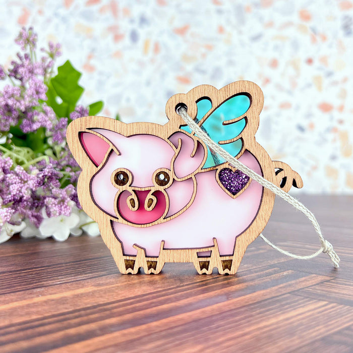 The When Pigs Fly Ornament, 3" by Forged Flare® is a charming wooden flying pig figurine designed as a pink pig with wings. It features a small purple glitter heart on its side and includes a braided linen string loop at the top for hanging. The background displays purple and white flowers set on a wooden surface against a light-colored patterned wall, making it the perfect farmhouse mantel decoration.