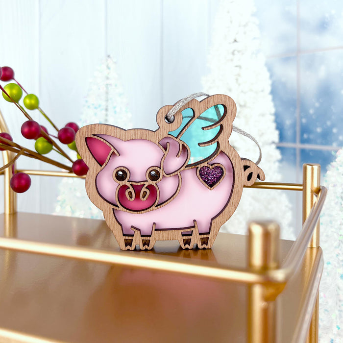 The "When Pigs Fly Ornament" from Forged Flare® features a charming wooden pig with a glittery purple heart and iridescent wings, displayed elegantly on a gold shelf. The festive background includes red berries and white Christmas trees, creating the perfect scene for holiday cheer. Ideal for farm decor enthusiasts or anyone who loves whimsical decorations!