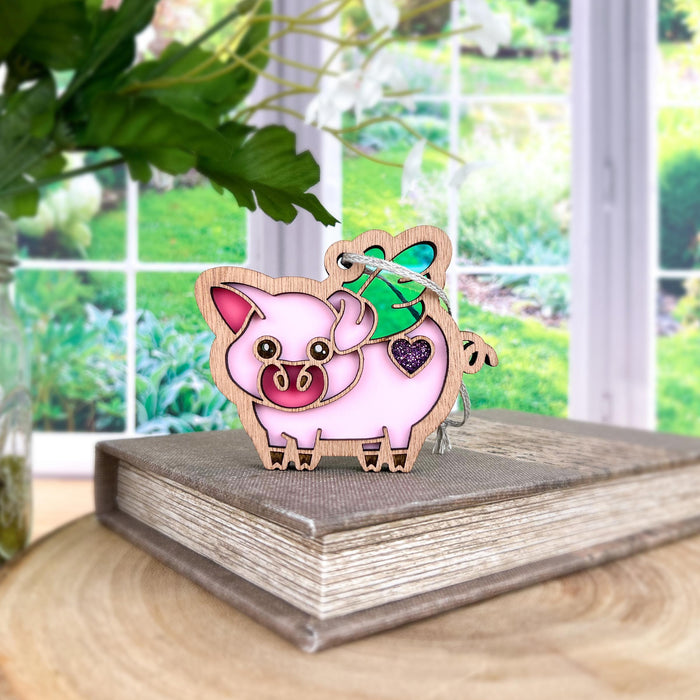 The whimsical When Pigs Fly Ornament, 3" from Forged Flare® features a charming pig with a pink body and wooden border, standing gracefully on a closed book. Adorned with iridescent wings and decorated with heart-shaped accents, this delightful piece is set against the backdrop of a bright window that opens up to reveal a lush green garden.