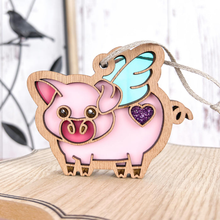 The Forged Flare® When Pigs Fly Ornament, 3", is displayed on a wooden surface. This adorable ornament features a pink pig with a blue wing and a purple heart on its side. The pig's snout is detailed in red, and it includes a string for easy hanging. The background showcases light wood walls with faint black bird decor, making it perfect farm decor!