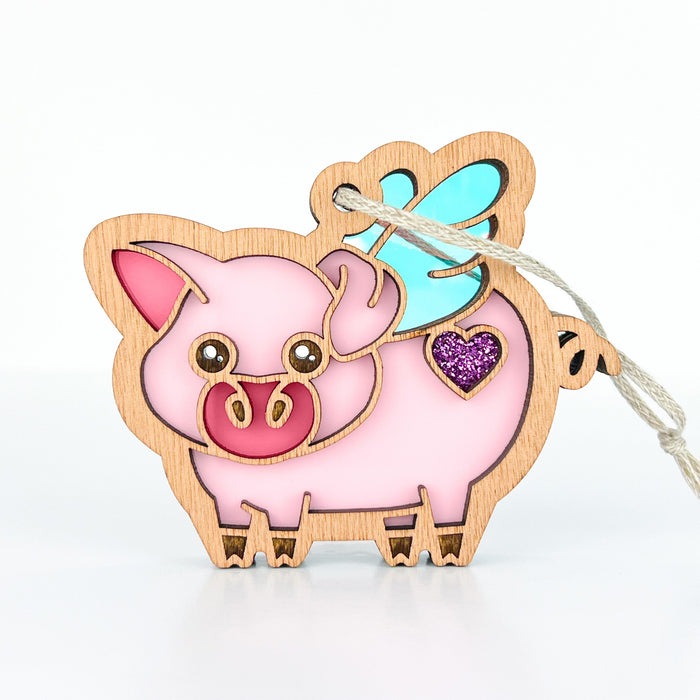 Add a touch of whimsy to your farm decor or farmhouse mantel with the "When Pigs Fly Ornament, 3"" by Forged Flare®. This charming wooden and pastel-colored stained glass–style ornament features a pink pig with blue wings, a pink snout, and a glittery purple heart on its side. It hangs delicately from a piece of braided linen string, making it an adorable addition to any space.