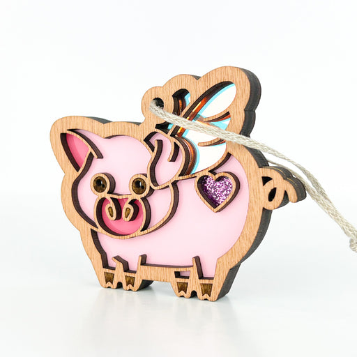 The When Pigs Fly Ornament, 3" by Forged Flare® is a decorative wooden flying pig adorned with pink and glittery purple details, featuring a heart-shaped accent and a iridescent wings on its back. This vibrant farmhouse mantel decoration includes intricate cutout designs and is equipped with a braided linen string at the top for easy hanging.