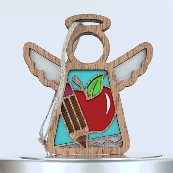 A wooden teacher ornament in the shape of an angel with wings. The angel has cut-out sections revealing colorful stained-glass-like details, including a red apple with a green leaf and a brown pencil. A braided linen string is attached for hanging.