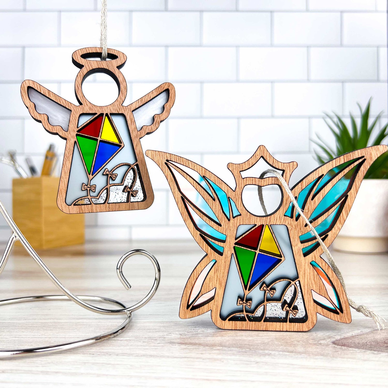 Two wooden Christmas ornaments shaped like an angel and a fairy are placed on a countertop with a white tiled background. Each ornament has colorful stained glass–style detailing. One hangs on a metal stand, while the other stands upright. A small green plant is visible on the right.